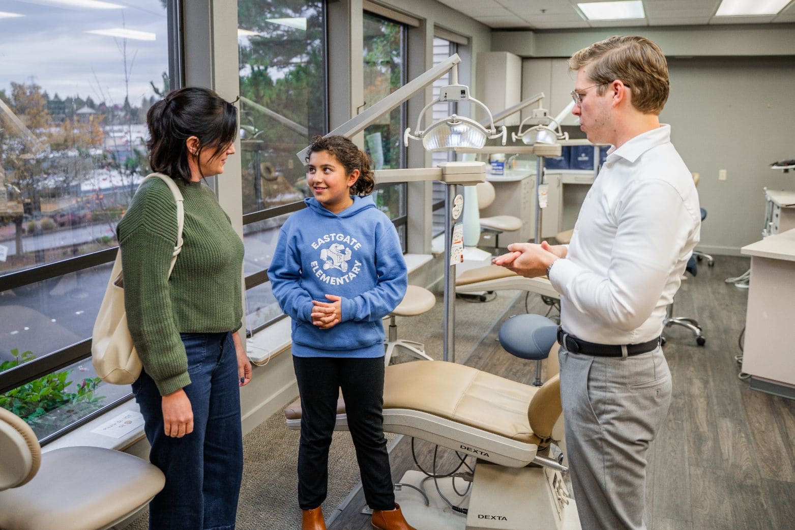 A woman and a child talk with a dentist in a dental clinic. The child is wearing a blue sweatshirt, the woman is in a green sweater, and the dentist is in a white shirt and gray pants.