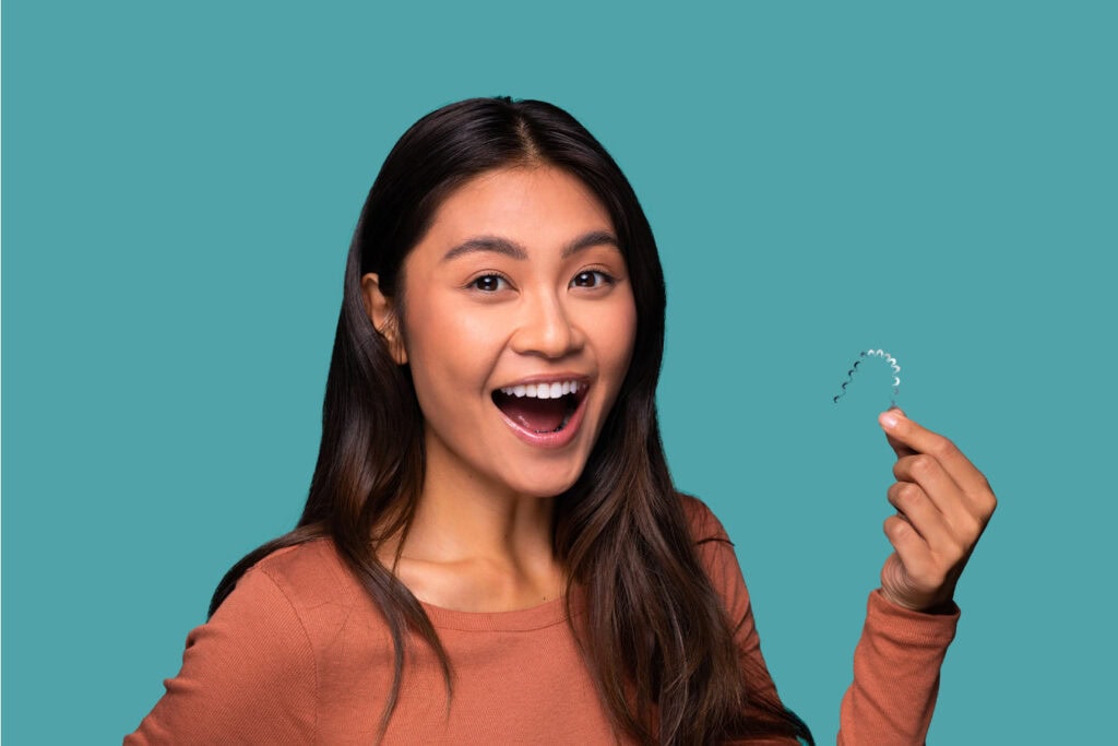 Young Asian woman smiling and holding an Invisalign aligner, isolated on a teal background.
